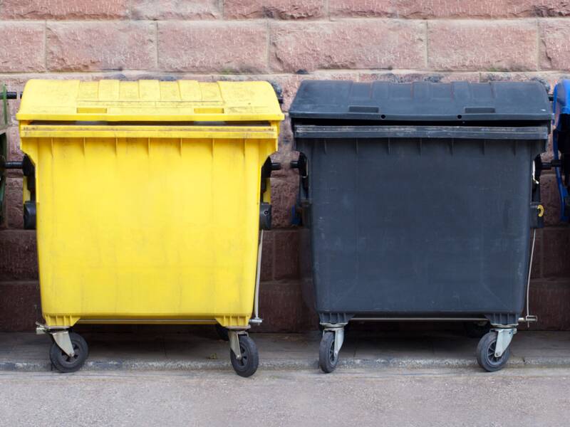 Grüner, gelber, schwarzer und blauer Müllcontainer nebeneinander vor einer Hauswand aus Backstein 