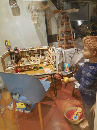 Ein Schaufenster mit einem Tisch voller Spielzeug, einer Pyramide mit Kerzen und einer Kinderpuppe in einem blauen Oberteil, die ein goldenes Objekt hält.