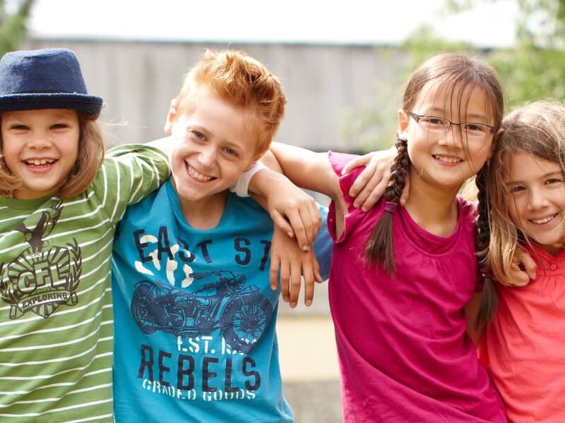 Sechs in die Kamera lächelnde Kinder stehen in einer Reihe nebeneinander und haben die Arme umeinander gelegt