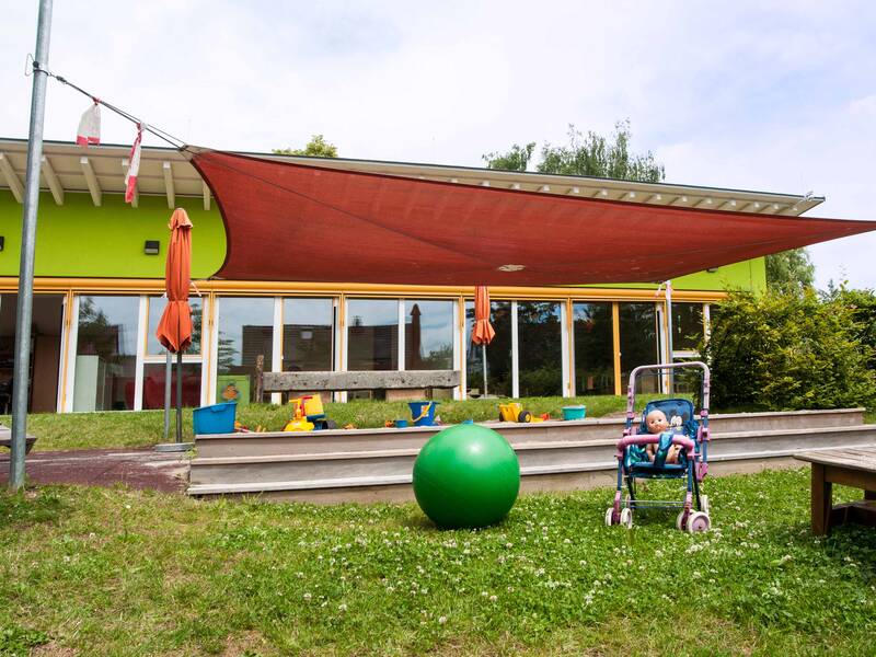 Sandkasten mit Sonnensegel im Garten der Evangelischen Kinderkrippe Zwergenstüble, davor ein Puppenbuggy mit einer Puppe und ein großer grüner Ball