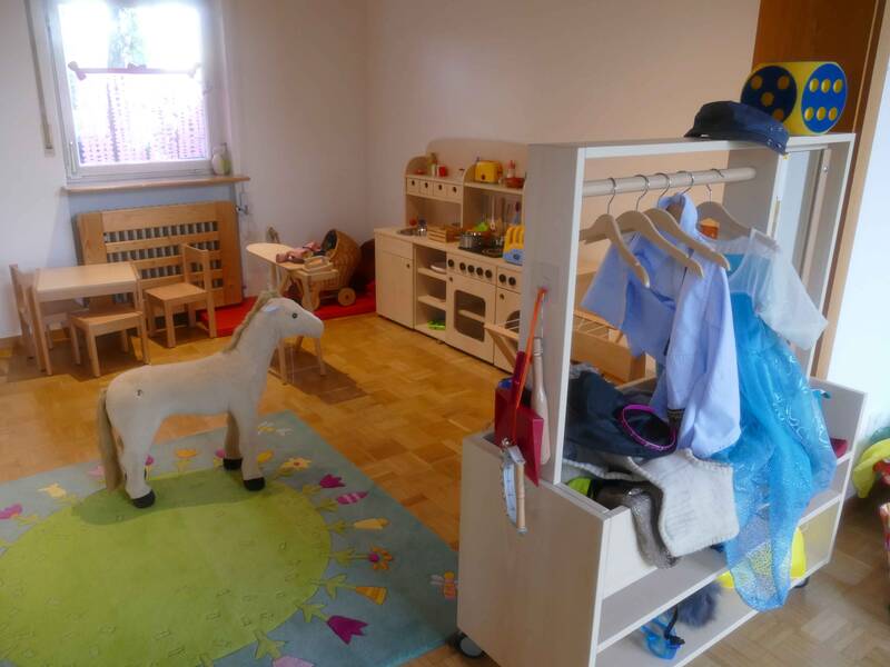 Gruppenraum im Kindergarten Ringweg in Roßwälden mit einem Spielpferd auf einem bunten Teppich und einem Kindertisch mit Stühlen, einer Spielküche aus Holz und einer Garderobe mit Kleidern zum Verkleiden. 