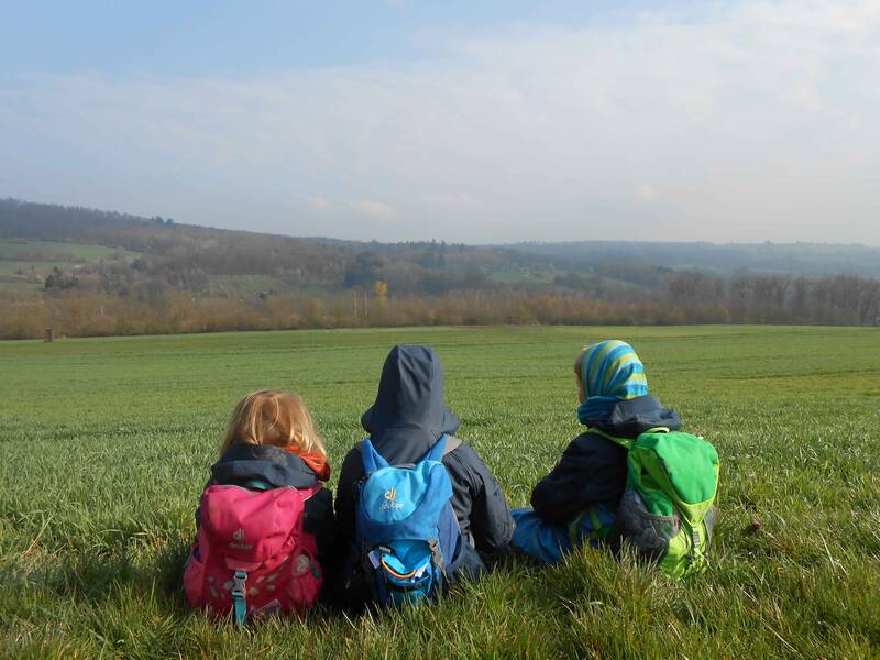 Drei Kinder des Rotmilane Naturkindergarten Weiler sitzen mit Rucksäcken auf einer Wiese und schauen auf Felder und Hügel