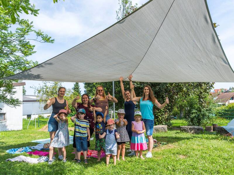 6 Kinder in bunter Sommerkleidung gemeinsam mit fünf Erzieherinnen unter einem grauen Sonnensegel im Garten des Kindergartens Pusteblume in Weiler.