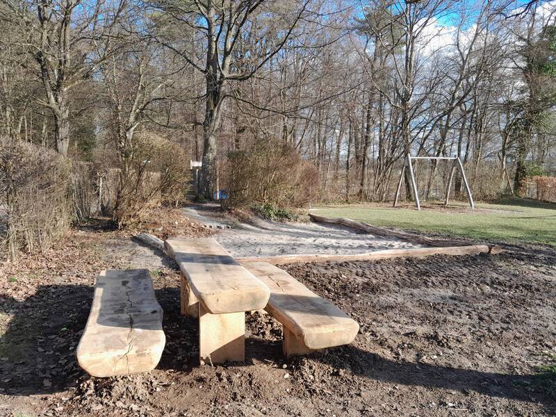 Sitzgruppe bestehend aus einem Tisch mit zwei Bänken aus Holz. Im Hintergrund eine Schaukel am Spielplatz Buschel