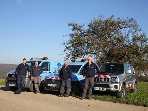 Vier Mitarbeiter der Stadtwerke stehen vor drei blauen Fahrzeugen der Stadtwerke, die an einem Feldweg vor einem Baum parken